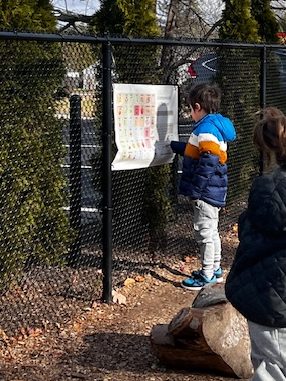 A child wearing a colorful jacket stands beside a fence, examining a poster attached to it, while another child is partially visible in the foreground.
